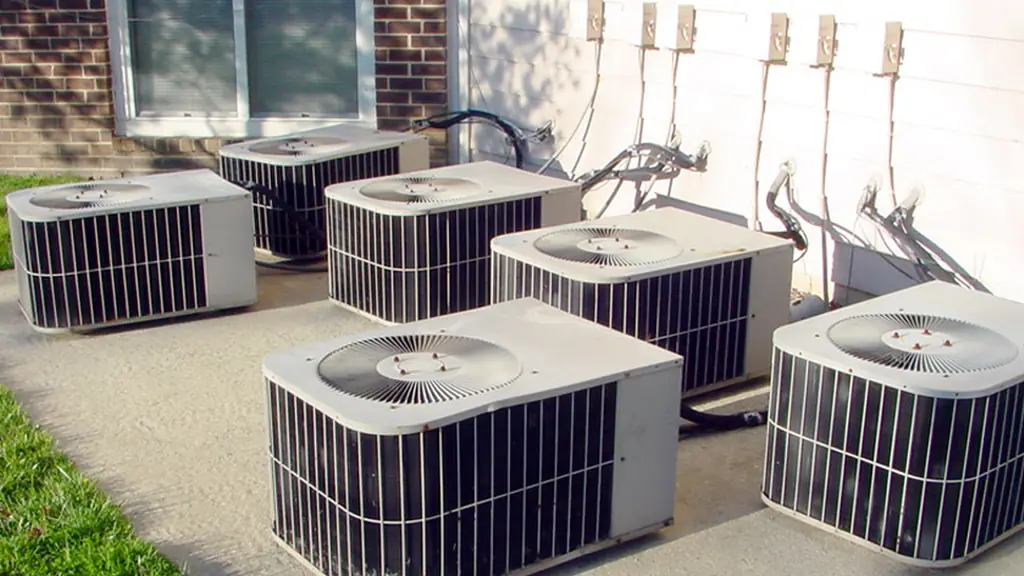 A row of six residential air conditioning condensers installed on a concrete pad along the side of a building